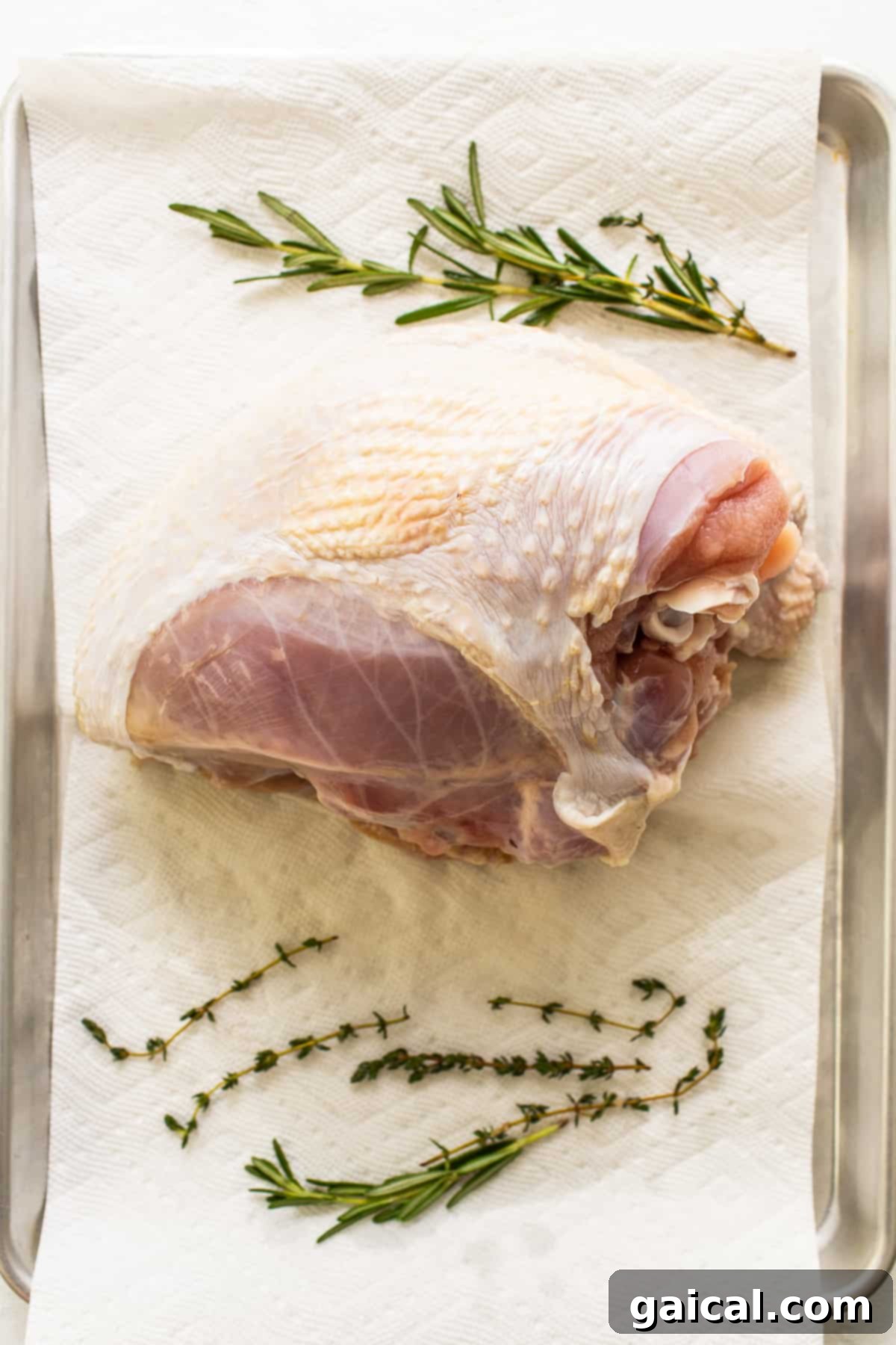 A turkey breast resting on a paper towel with fresh herbs in the background, ready for seasoning.