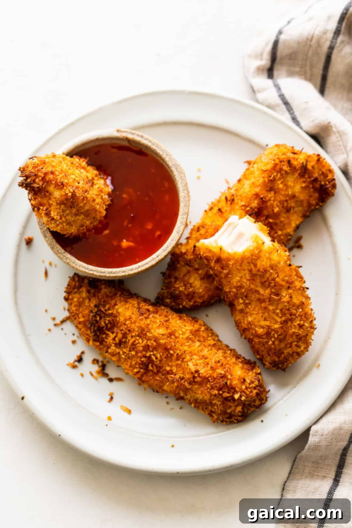 A plate of golden-brown, crispy coconut chicken tenders served with a side of dipping sauce, garnished with fresh cilantro.