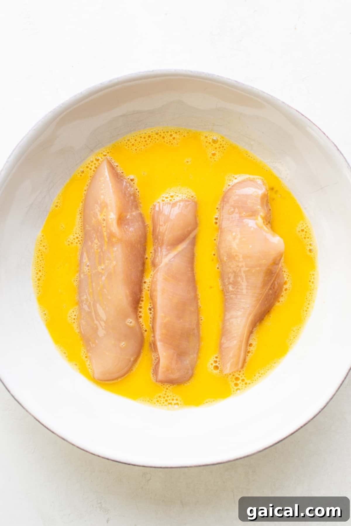 Raw chicken tenders being dipped in an egg wash in a bowl, preparing for breading.