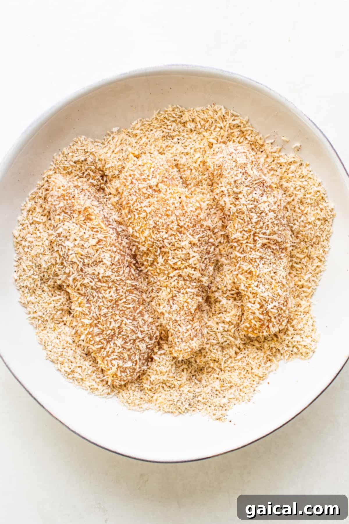 Raw chicken tenders thoroughly coated in a vibrant mixture of shredded coconut, breadcrumbs, and spices, ready for cooking.