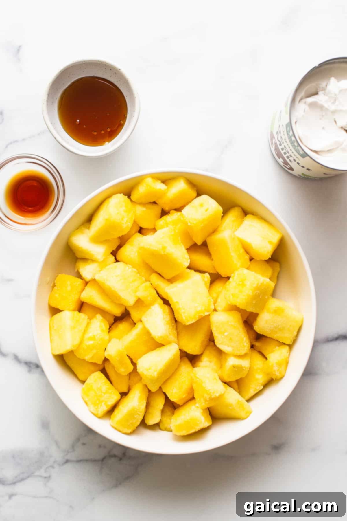 Essential Ingredients for Dole Whip Four bowls filled with the ingredients for Dole Whip: frozen pineapple chunks, a can of full-fat coconut milk, a bottle of vanilla extract, and a jar of honey, arranged on a kitchen counter.
