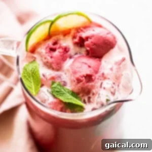 A large glass pitcher filled with vibrant raspberry vodka punch, with ginger beer being poured in and scoops of sorbet added for a frosty finish.