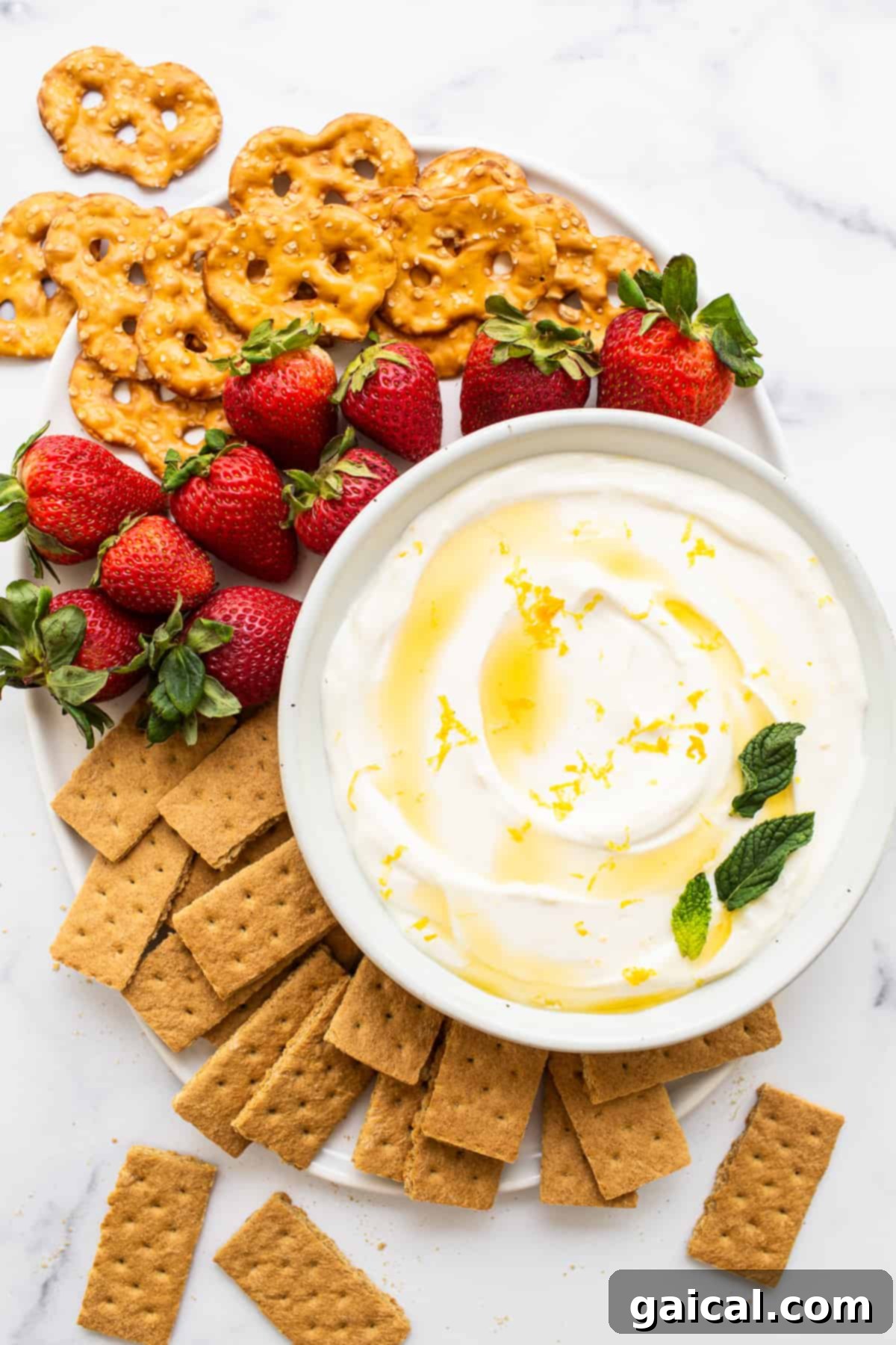 A vibrant bowl of creamy fruit dip is nestled amongst a colorful bounty of fresh berries, crunchy pretzels, and golden graham crackers on a rustic wooden board, illustrating perfect party presentation.