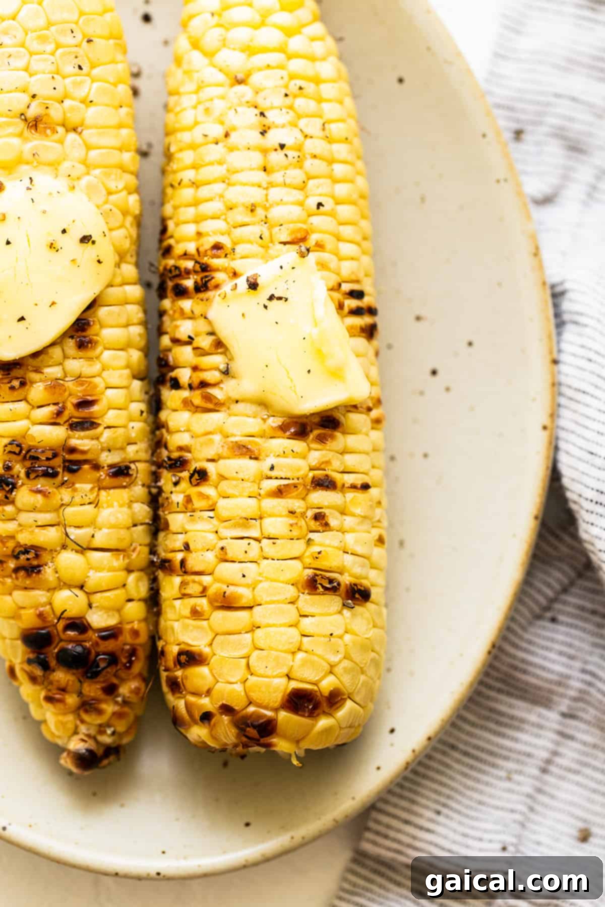 Close-up of grilled corn on the cob topped with melted butter, salt, and pepper, resting on a clean white serving plate, ready to be enjoyed.