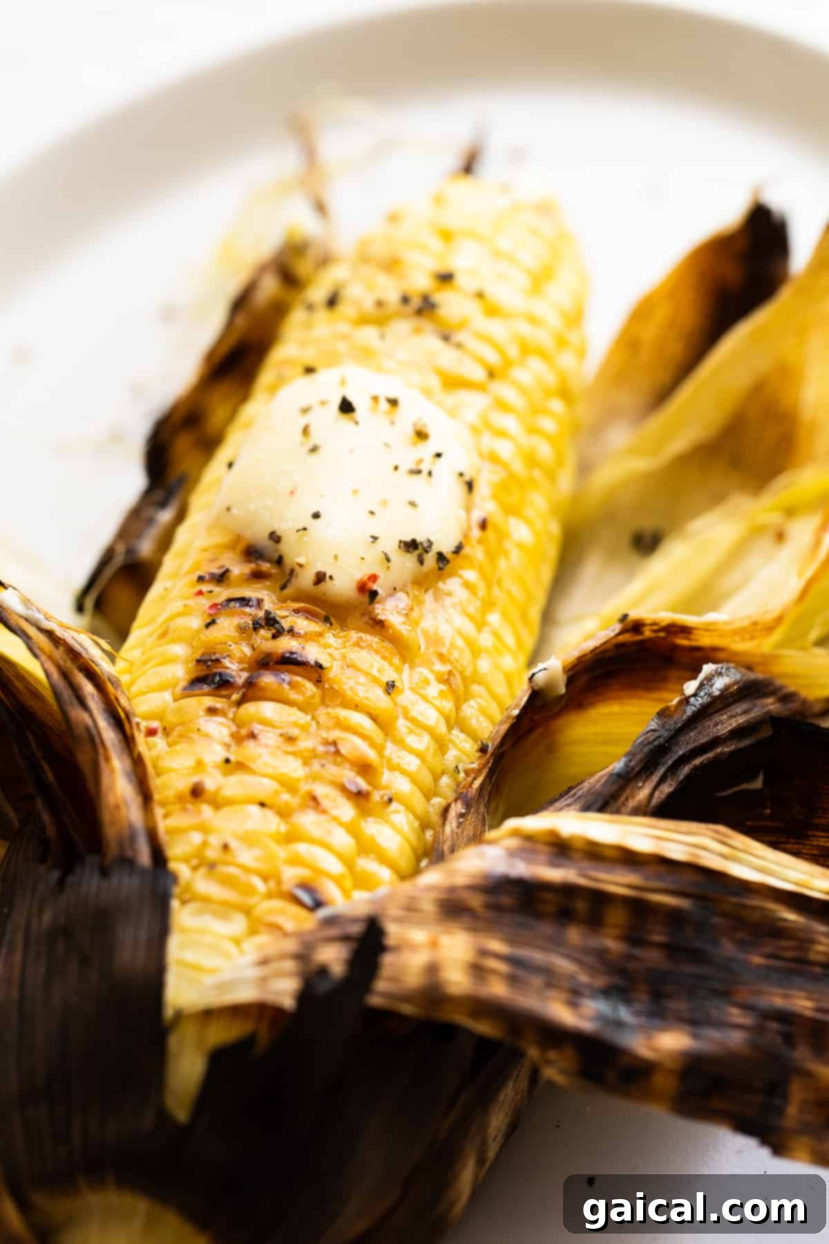 Three golden ears of grilled corn on the cob, generously buttered and seasoned with coarse salt and freshly ground black pepper, displayed on a wooden surface.