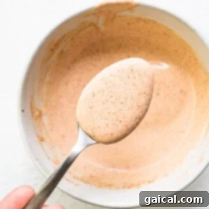 A person holding a spoon full of a creamy brown burger sauce in a bowl.
