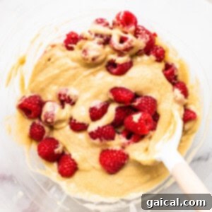Close-up of muffin batter in a bowl, generously speckled with fresh raspberries, ready to be scooped.