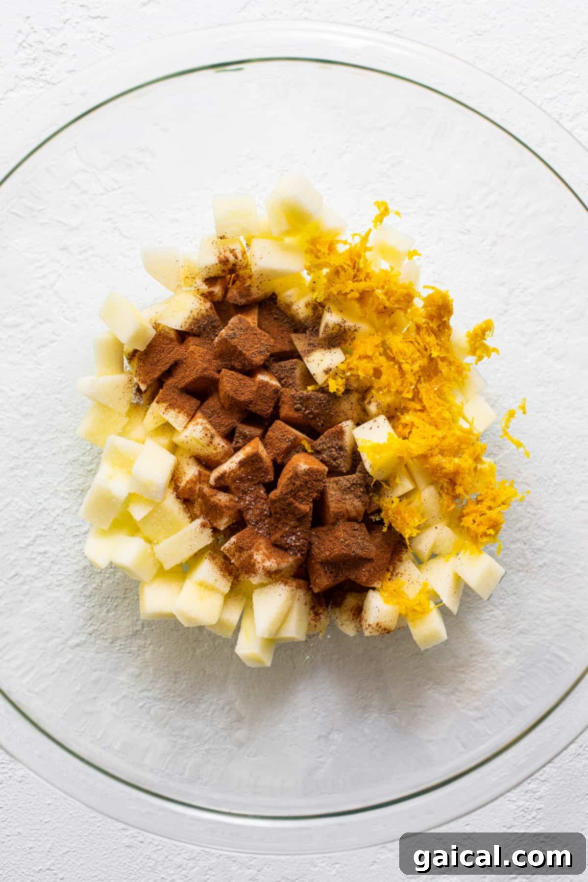 Mini apple crisps are a fun way to serve traditional apple crisps but in a single serving. Top them with vanilla ice cream for the perfect fall dessert. Close-up view of the raw apple crisp filling in a mixing bowl, showcasing diced apples coated in vibrant orange zest and spices.