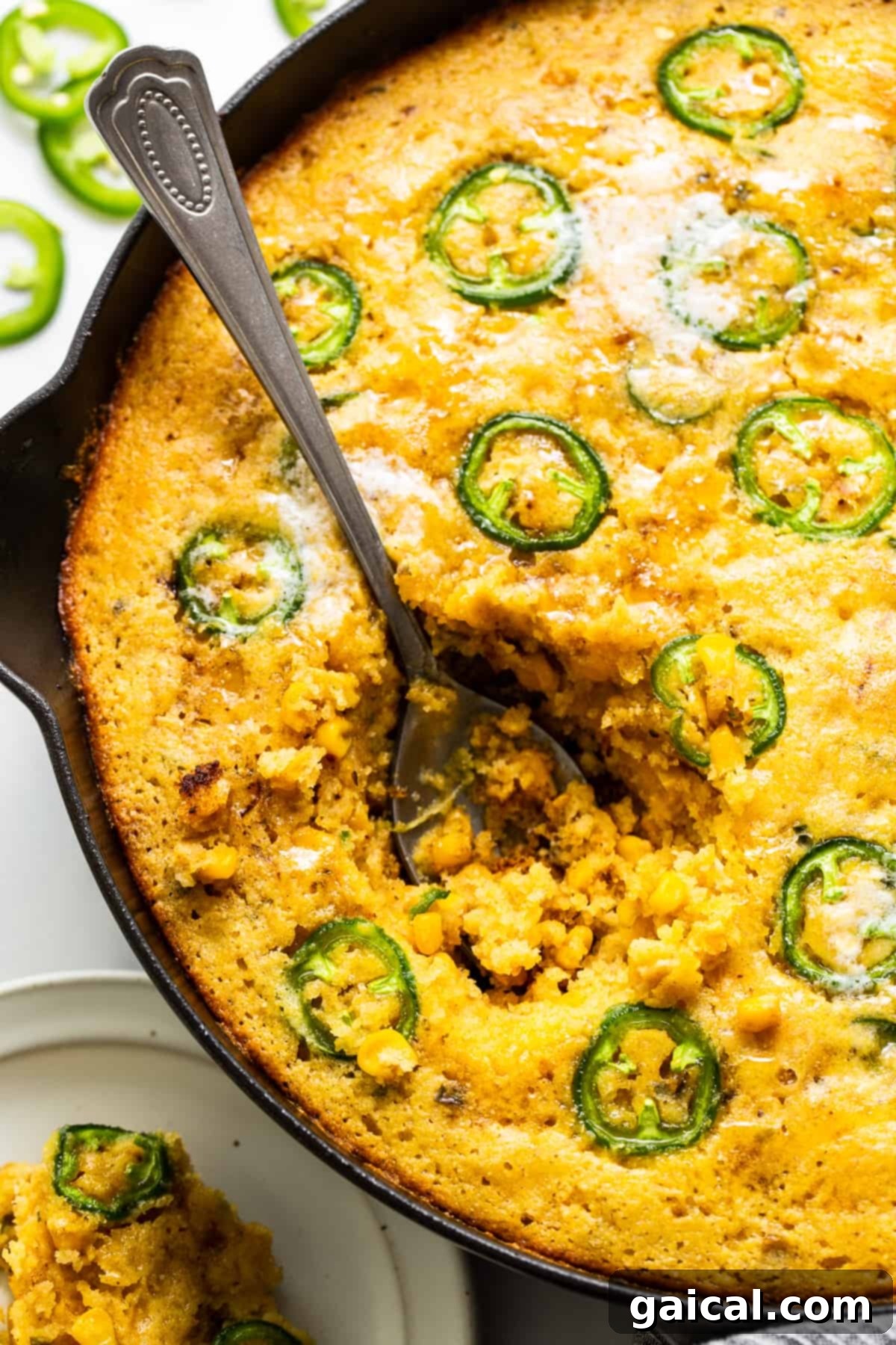 This jalapeño corn pudding recipe is made completely in a cast iron skillet in under an hour. It packs the perfect combo of sweet and spicy that you'll absolutely love! A large serving spoon scooping a generous portion of creamy jalapeño corn pudding from a hot cast iron skillet, showcasing its soft, delicious texture.