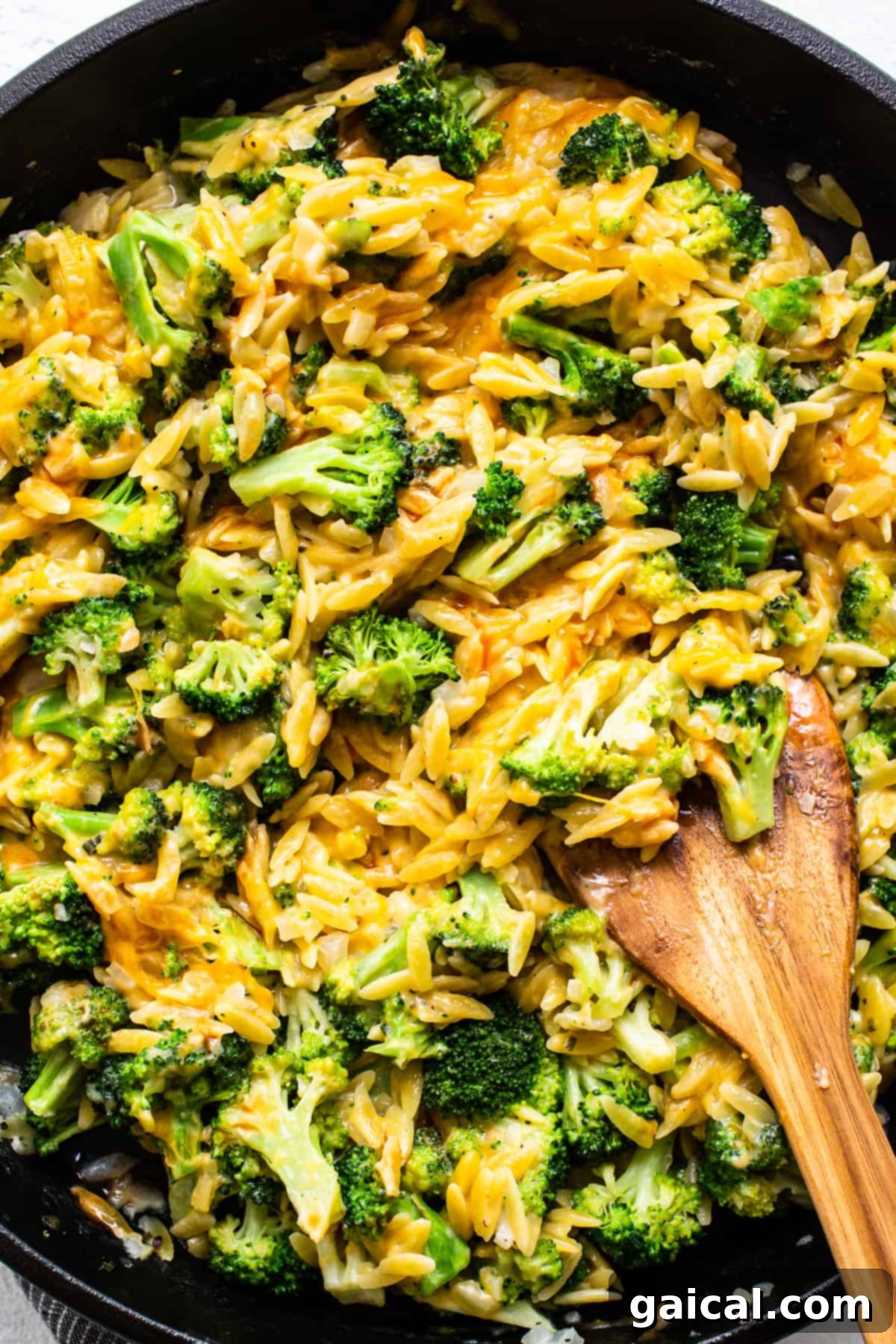 Mixing Ingredients for a Harmonious Casserole A close-up of a chef mixing creamy orzo and bright green broccoli with melted cheddar cheese in a cast iron skillet, ensuring all ingredients are well combined.