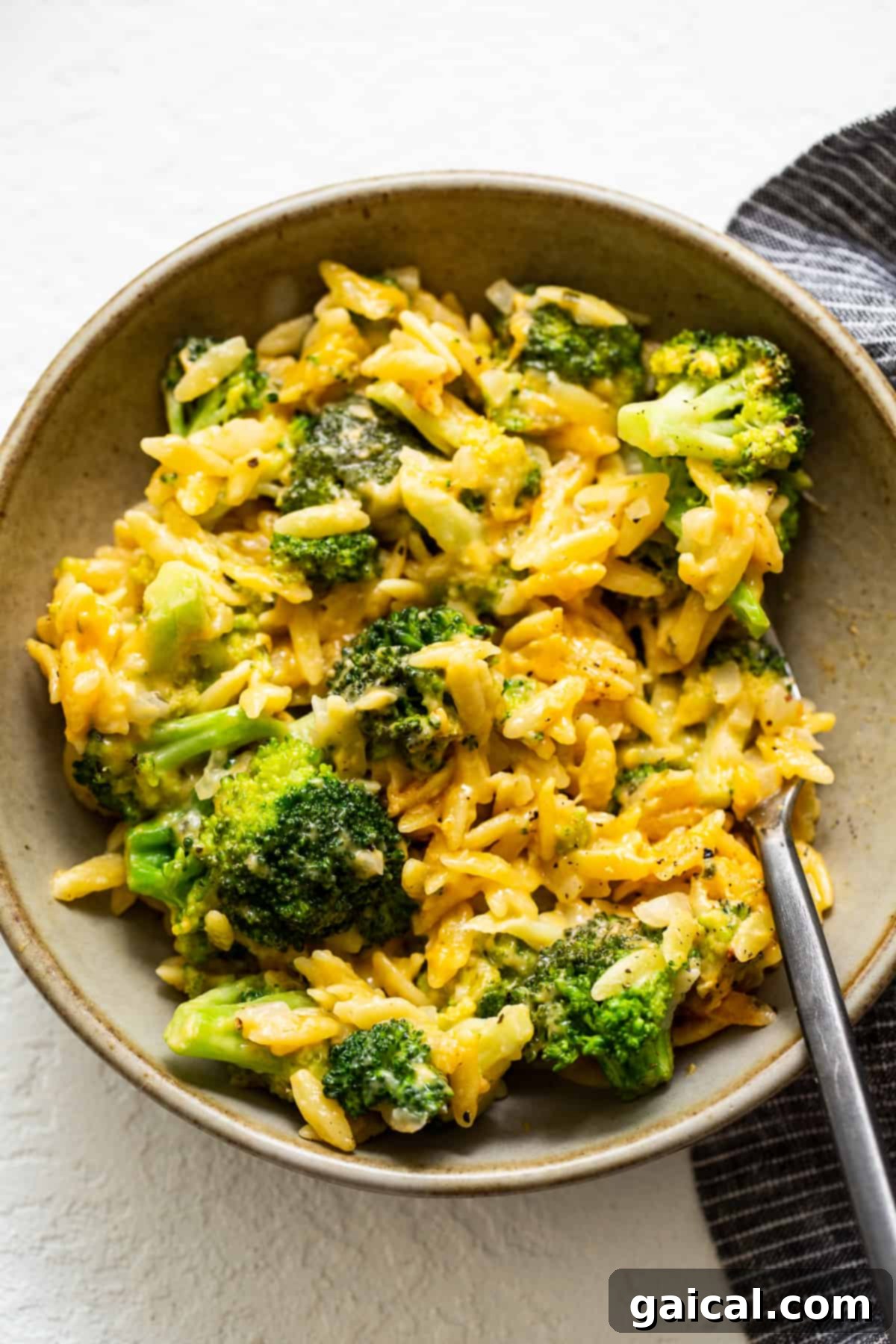 Broccoli Cheese Casserole Ready to Enjoy A serving of creamy broccoli cheese orzo casserole in a white bowl, garnished with fresh herbs and a fork resting beside it, ready to be eaten.