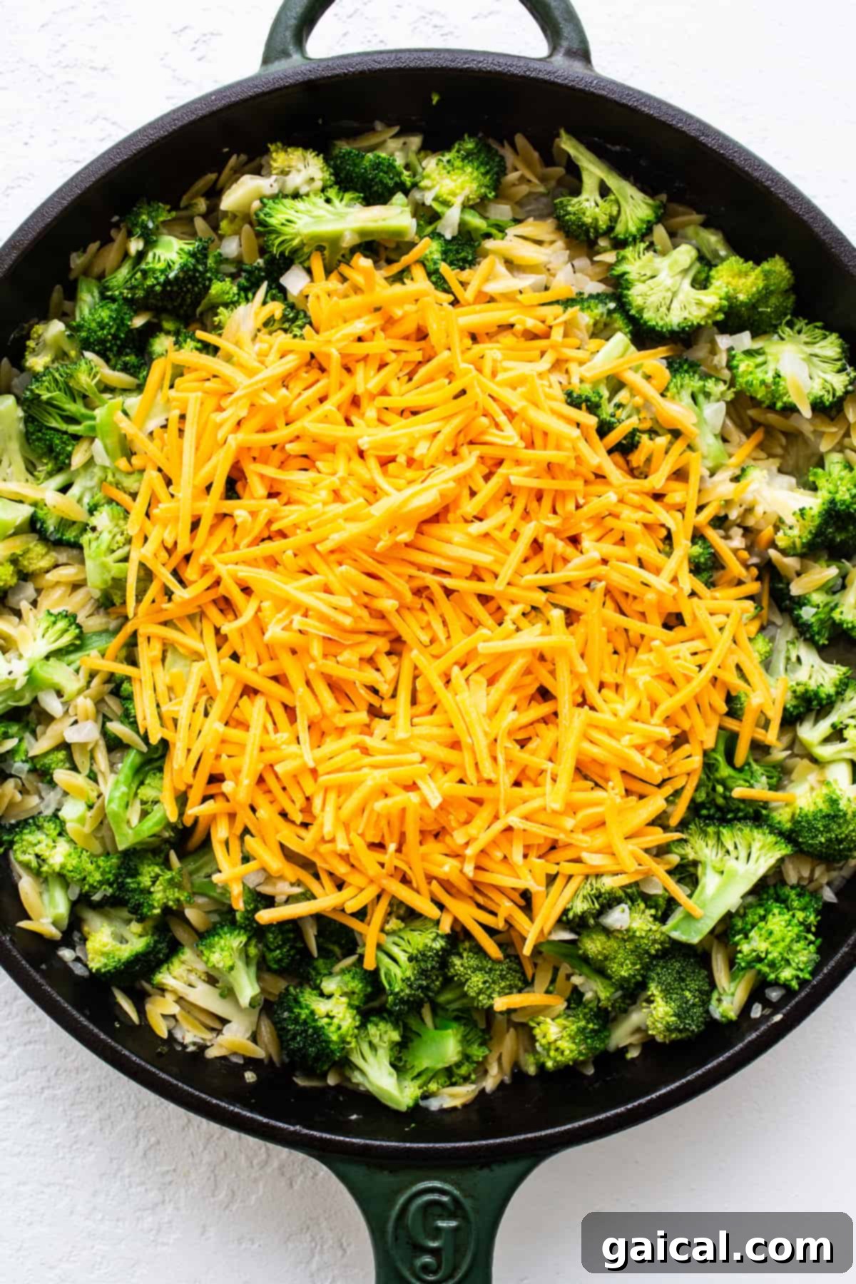 Cheese Melting into the Casserole Shredded cheddar cheese melting over cooked orzo and vibrant green broccoli florets in a cast iron skillet.