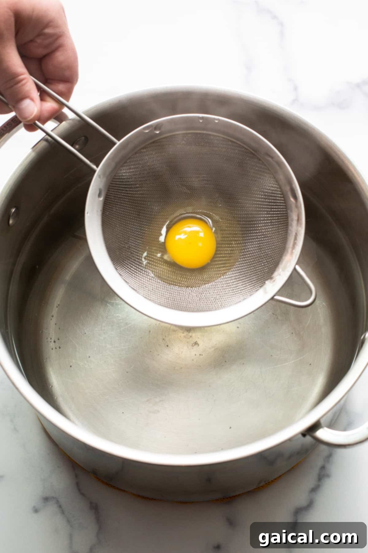 Gentle placement of a strained egg into simmering water for poaching. A raw egg in a mesh strainer being carefully placed into a pot of simmering water.