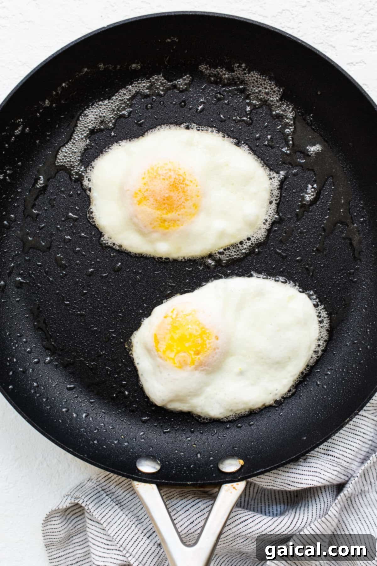 Two over hard eggs frying in a skillet with melted butter, the whites fully opaque and the yolks visibly firming up.