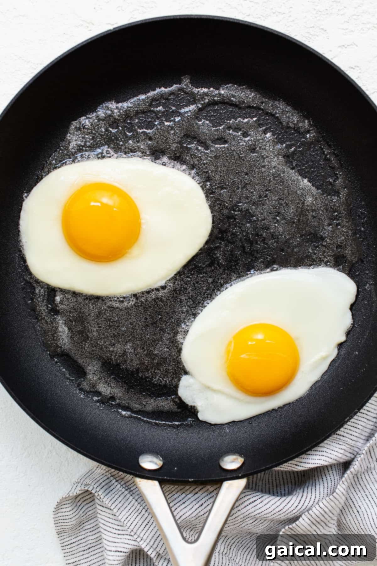Two over hard eggs continuing to cook gently in a non-stick skillet, their whites fully set and yolks solidifying.