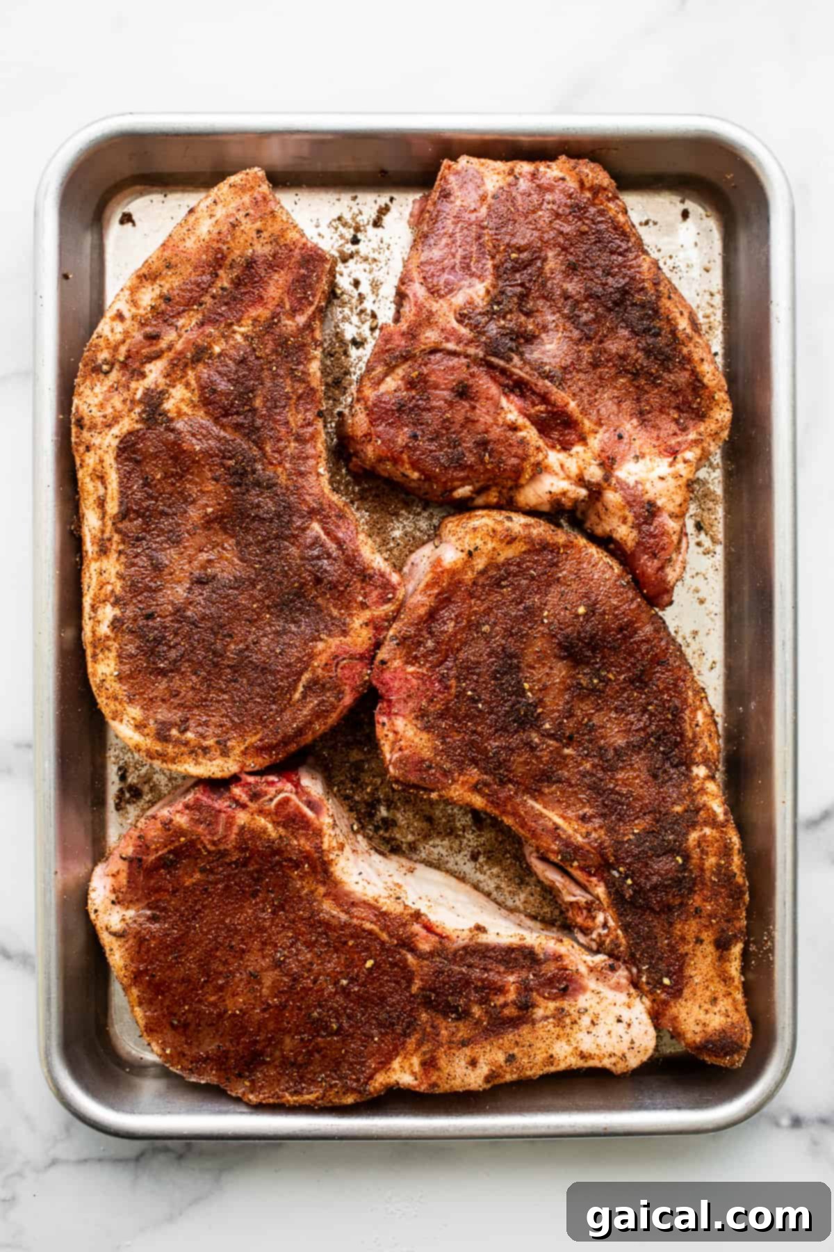 Seasoned Raw Pork Chops Four raw, thick-cut bone-in pork chops on a baking sheet, seasoned with a dry rub.