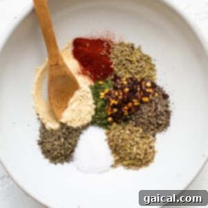 A small white bowl filled with a variety of dried spices, including paprika, thyme, rosemary, parsley, oregano, and red pepper flakes, with a wooden spoon.