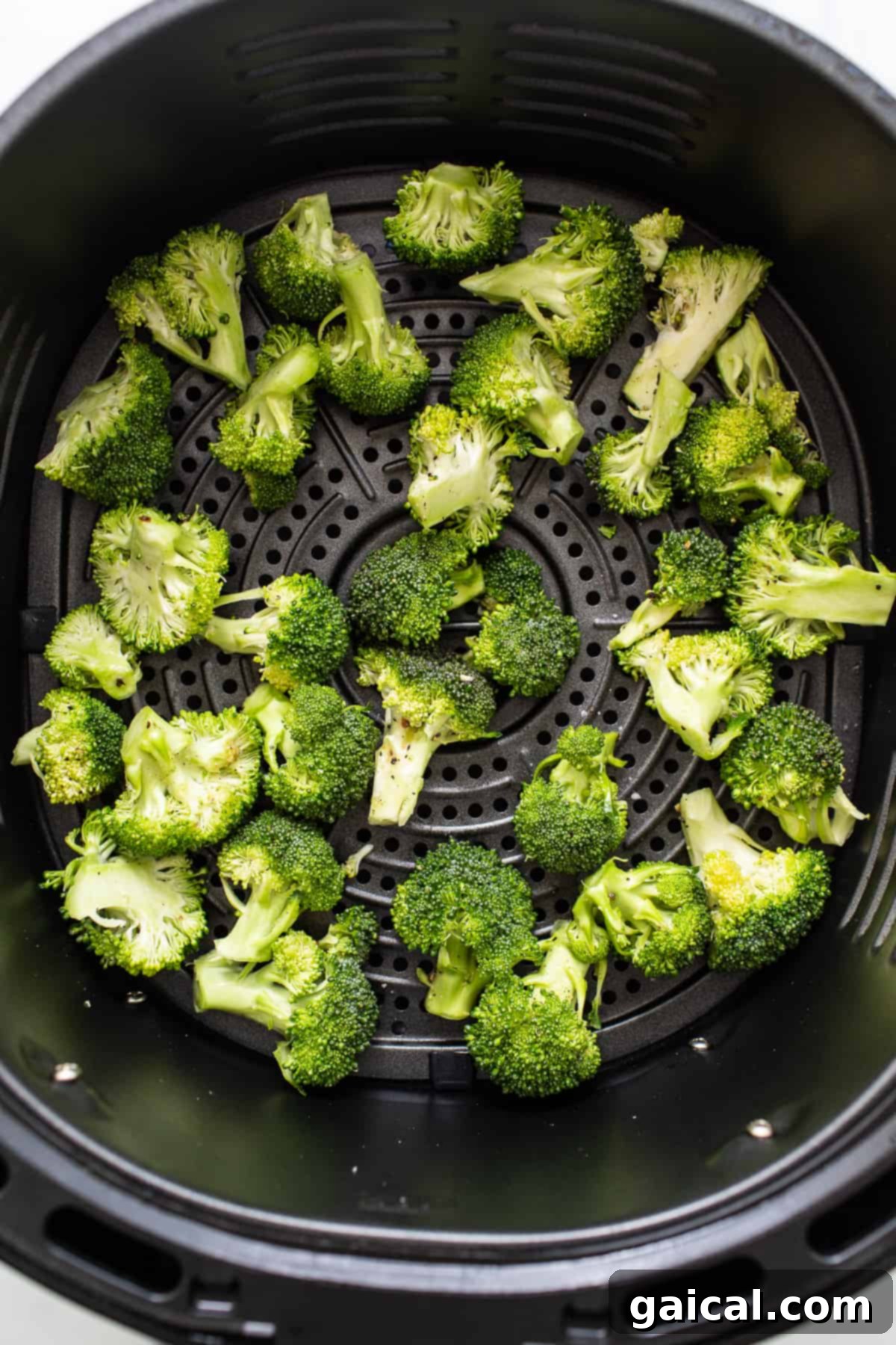Broccoli florets spread out evenly in the basket of an air fryer, beginning to crisp.