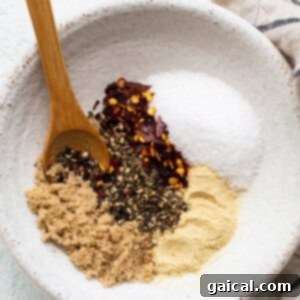 A bowl holds a savory steak seasoning mix of brown sugar, black pepper, crushed red pepper flakes, salt, and garlic powder, with a wooden spoon resting on the side.