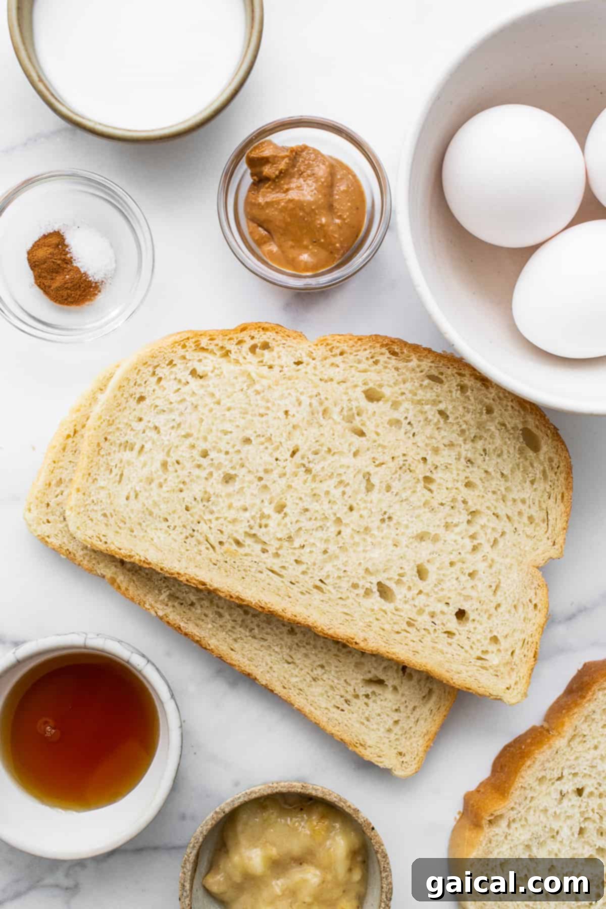 A collection of fresh ingredients laid out on a surface, including eggs, milk, bananas, maple syrup, cinnamon, peanut butter, bread, chocolate, and walnuts, ready for making air fryer chunky monkey french toast.