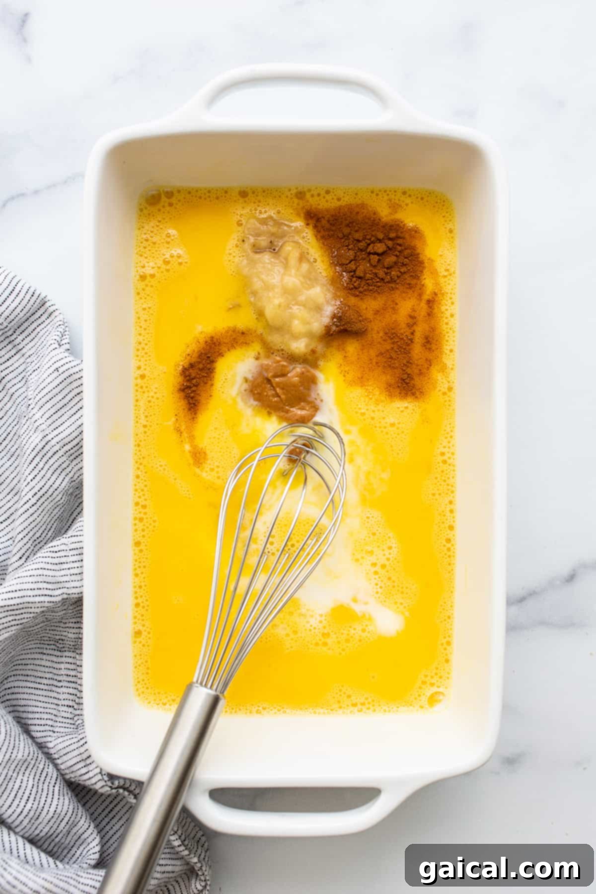 A close-up shot of a whisk swirling a creamy egg wash mixture, containing mashed banana, maple syrup, cinnamon, and peanut butter, in a white bowl.