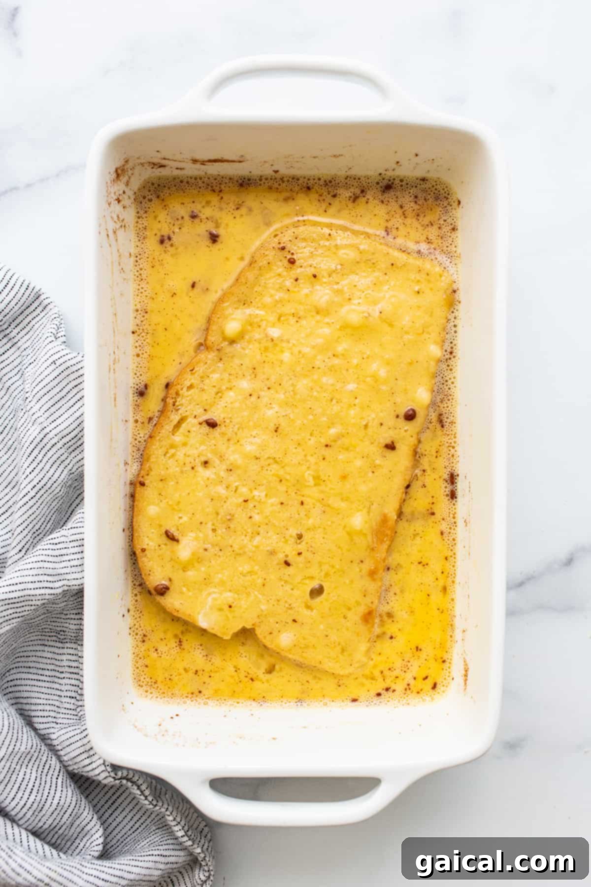 Two slices of thick bread are submerged in a creamy egg wash, absorbing the flavorful mixture in a white baking dish, preparing for air frying.