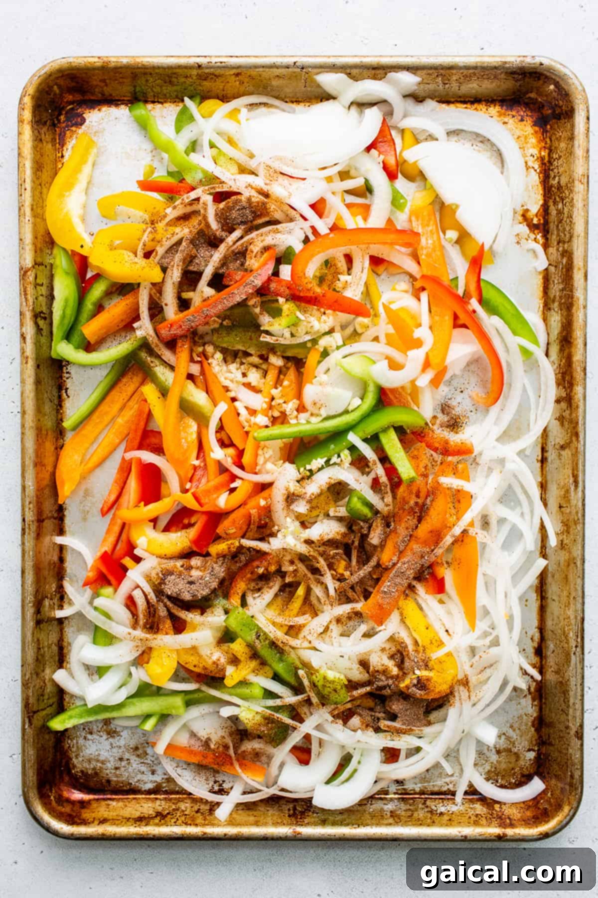 Uncooked sliced bell peppers and onions evenly spread on a baking sheet, seasoned and ready for roasting.