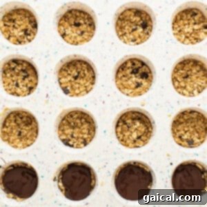 A muffin tray partially filled with granola bar bases and melted chocolate chips, showing the assembly of layers.