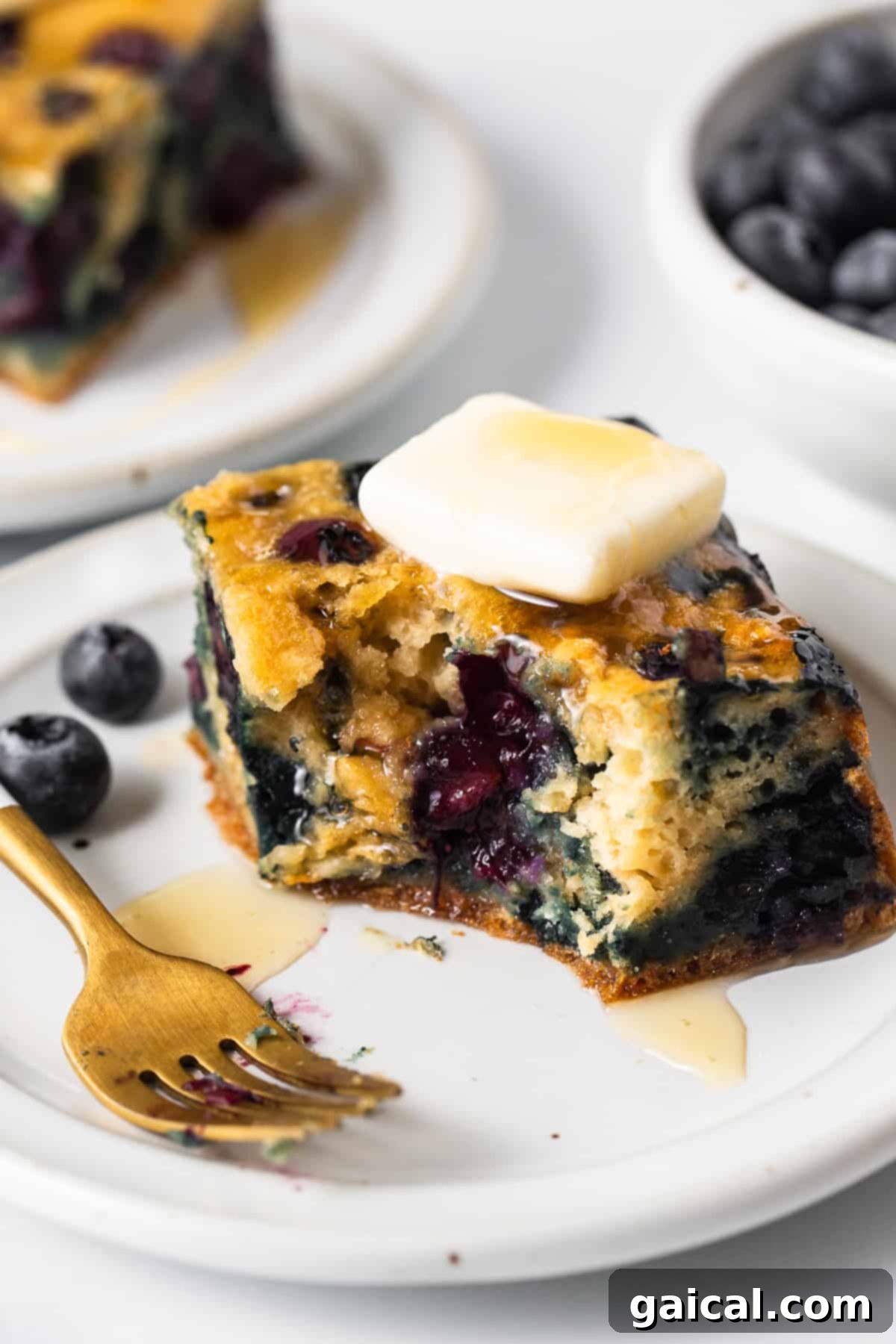 Golden Blueberry Coffee Cake 2 A beautifully baked slice of blueberry breakfast cake on a white plate, garnished with fresh blueberries and a light dusting of powdered sugar.