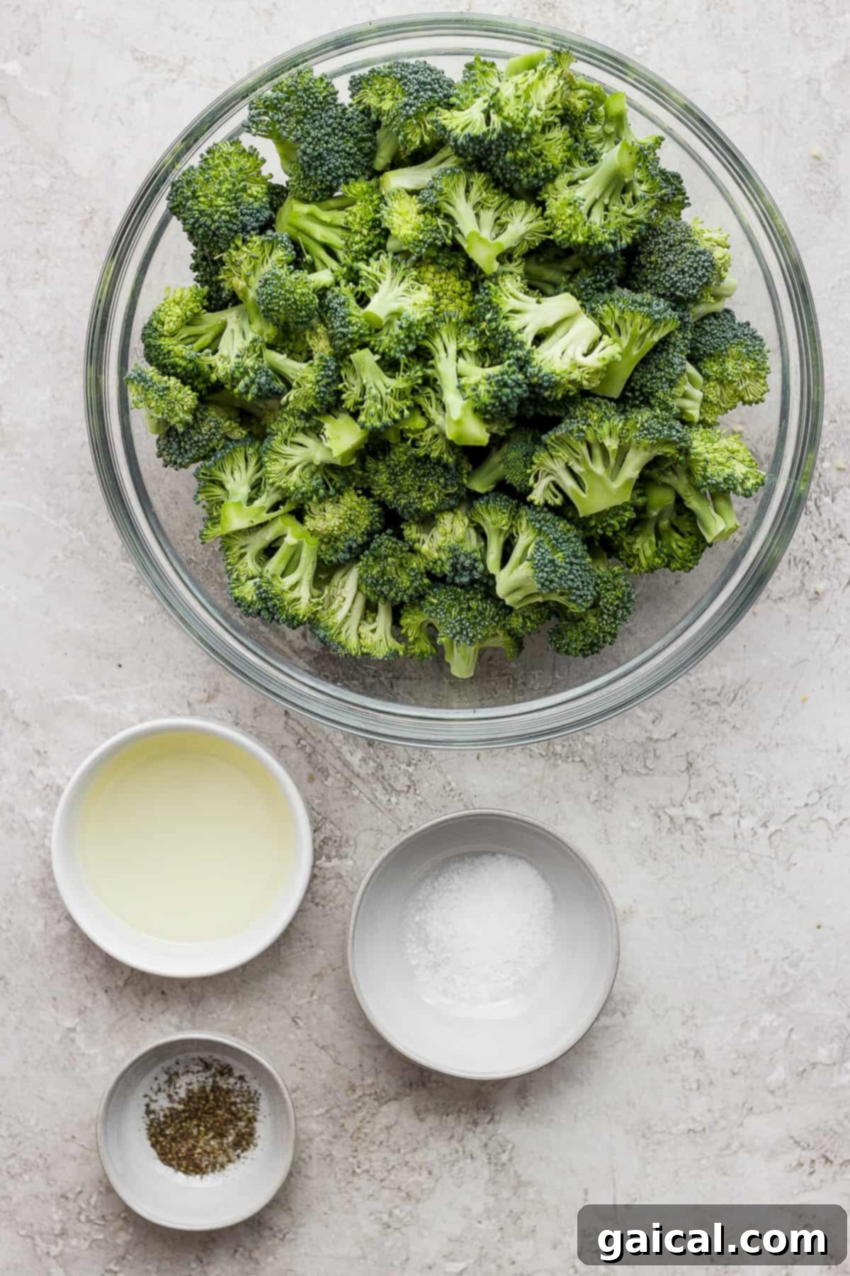 Charred Broccoli Perfection 3 A vibrant green bowl of fresh broccoli florets sits beside two smaller bowls, one holding olive oil and the other salt and pepper, highlighting the simple, essential ingredients for grilled broccoli.
