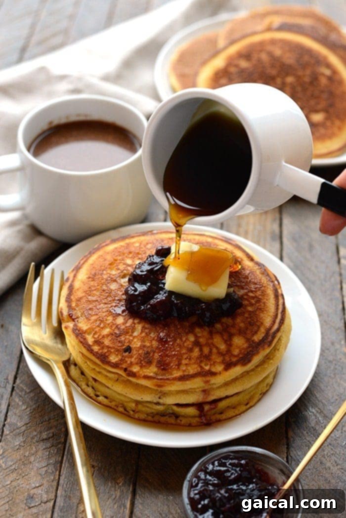 A stack of golden brown whole wheat cornmeal pancakes on a white plate, drizzled with maple syrup and garnished with fresh berries.