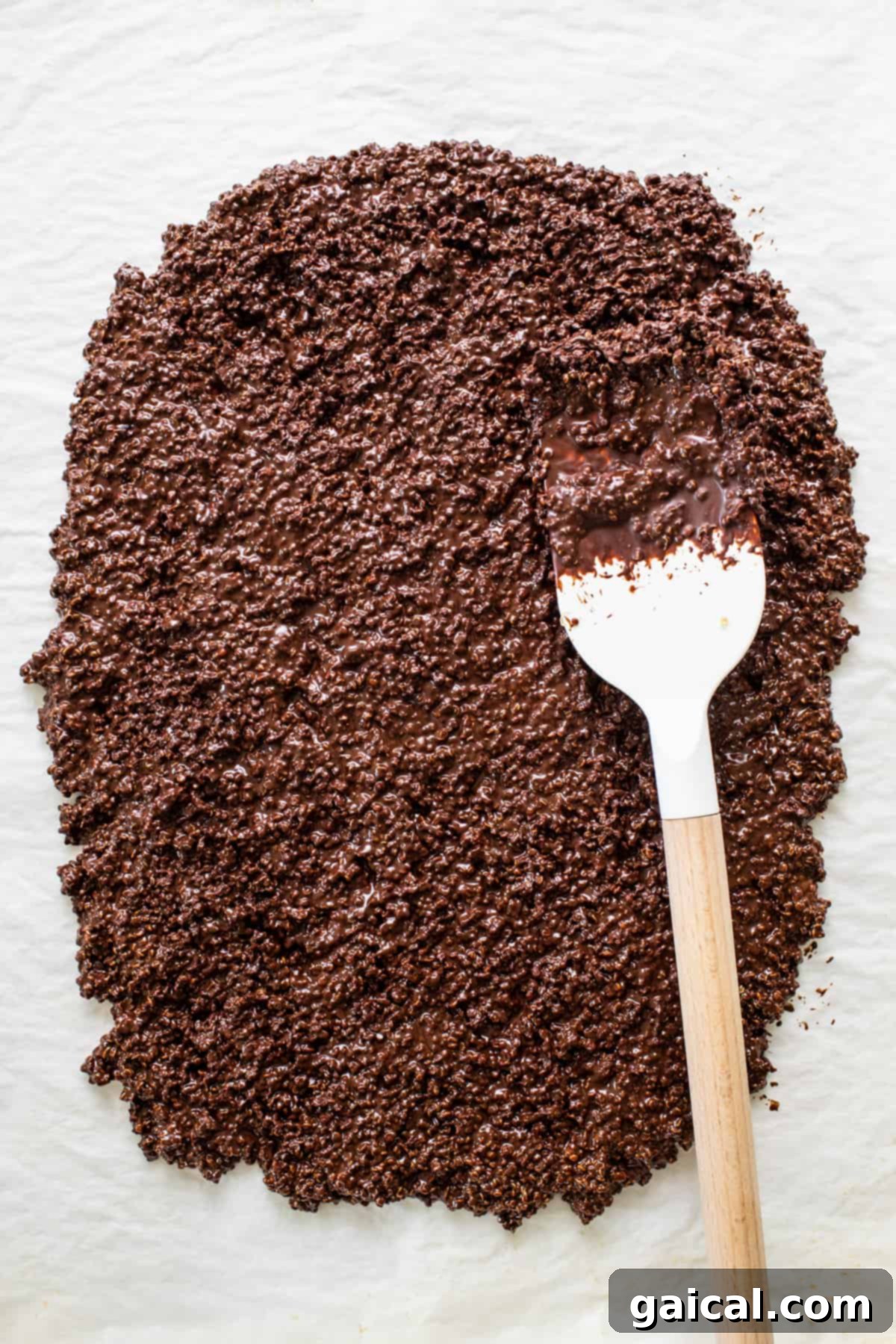 A spoon coated in glossy melted chocolate, poised to be incorporated into the crispy quinoa mixture, illustrating a key step in the recipe.