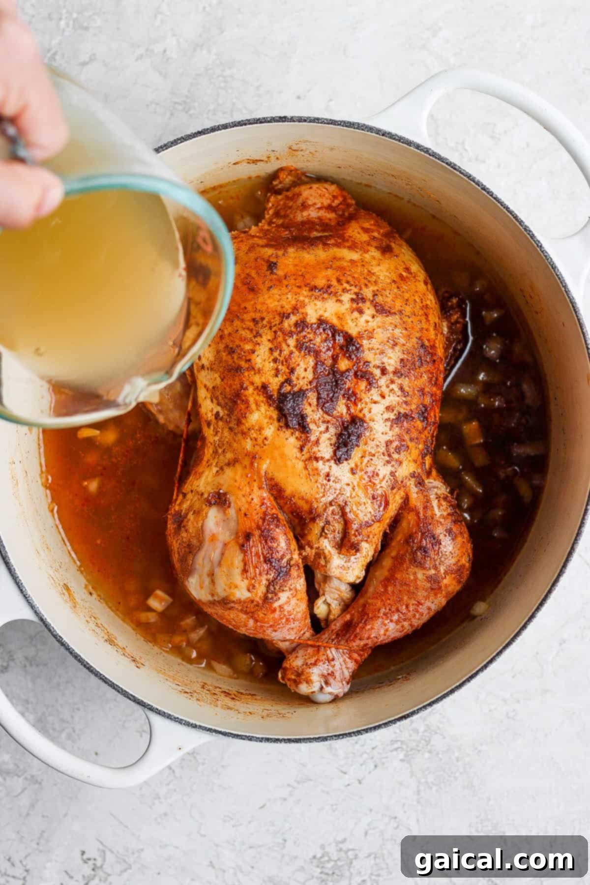 One-Pot Spanish Roast Chicken Dinner 6 A person carefully pouring broth over a seasoned whole chicken in a Dutch oven.