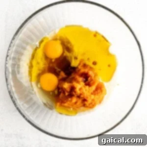 Cozy Pumpkin Spice Muffins 5 Two fresh eggs being whisked vigorously in a clear glass bowl, amidst other wet ingredients on a clean white background.