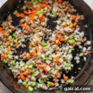 A cast iron skillet filled with browned Italian sausage, onions, garlic, carrots, and celery.