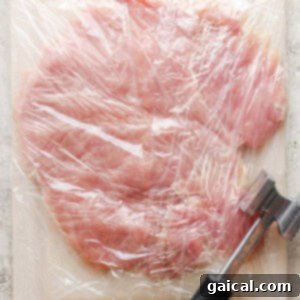 A butterflied turkey breast covered with plastic wrap, flattened on a cutting board.