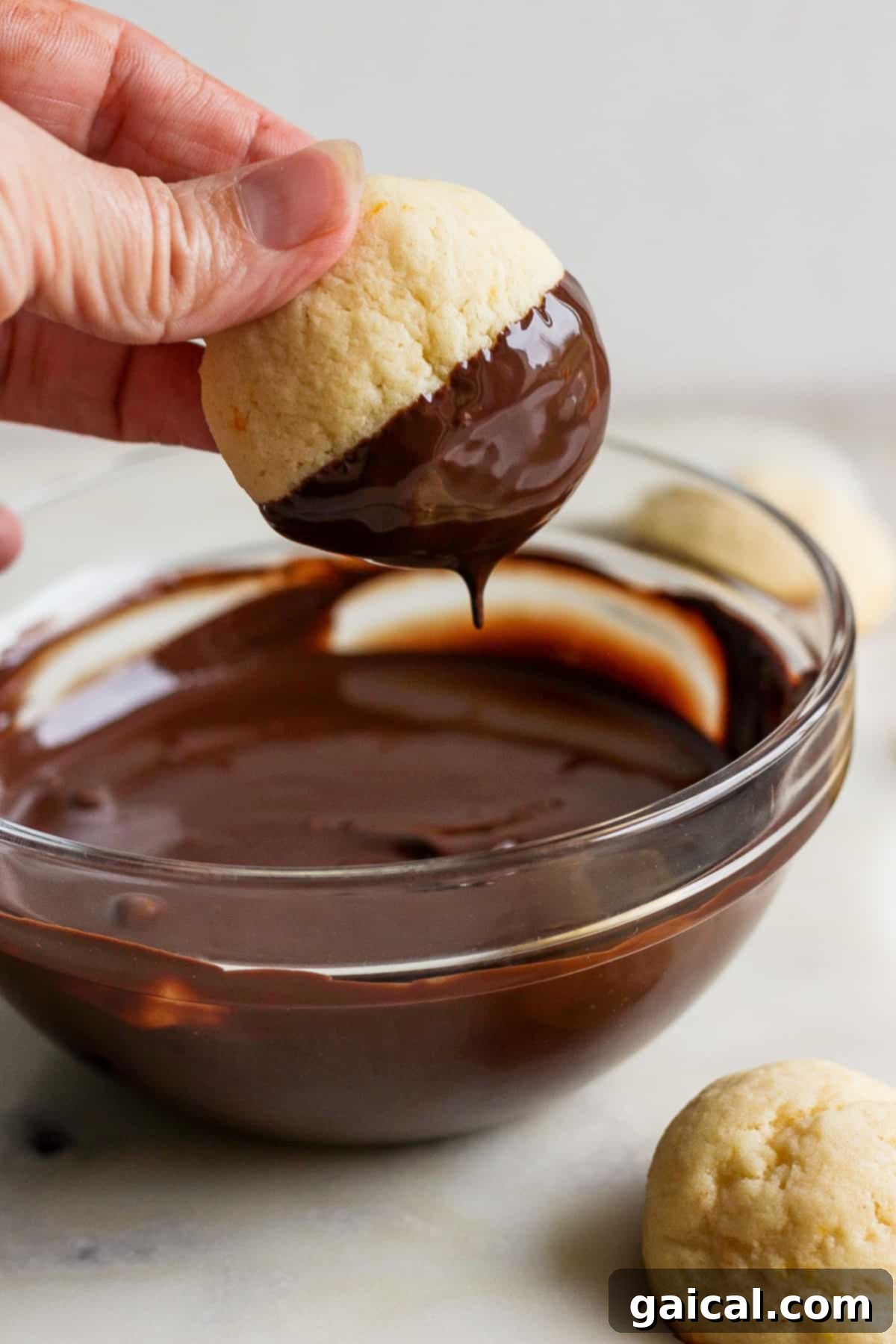 A person carefully dipping a freshly baked ricotta cookie into a bowl of melted dark chocolate, preparing it for its final flourish.