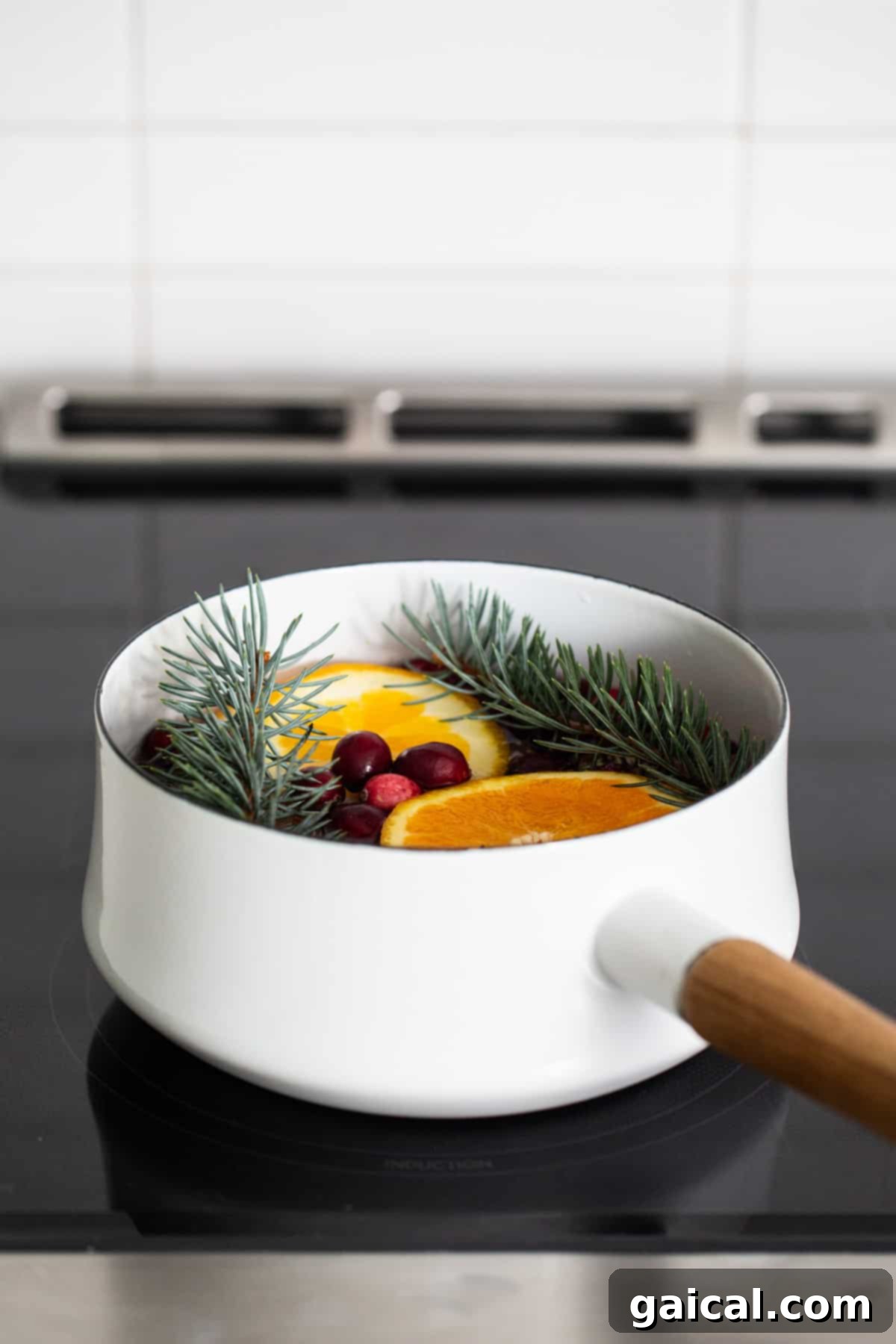 Cozy Holiday Aroma Pot 4 A simple white saucepan sitting on a stovetop, gently simmering with bright orange slices and fresh pine needles, filling the kitchen with a warm, natural scent.