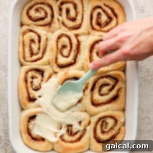 Nutty Brown Butter Banana Bread Cinnamon Rolls 13 A hand spreading the luscious brown butter cream cheese frosting over warm, baked cinnamon rolls in a dish.