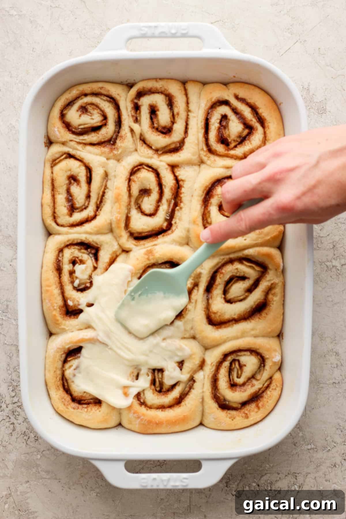 Nutty Brown Butter Banana Bread Cinnamon Rolls 5 A hand gently dipping a spatula into a dish of warm, iced cinnamon rolls, ready to be served.