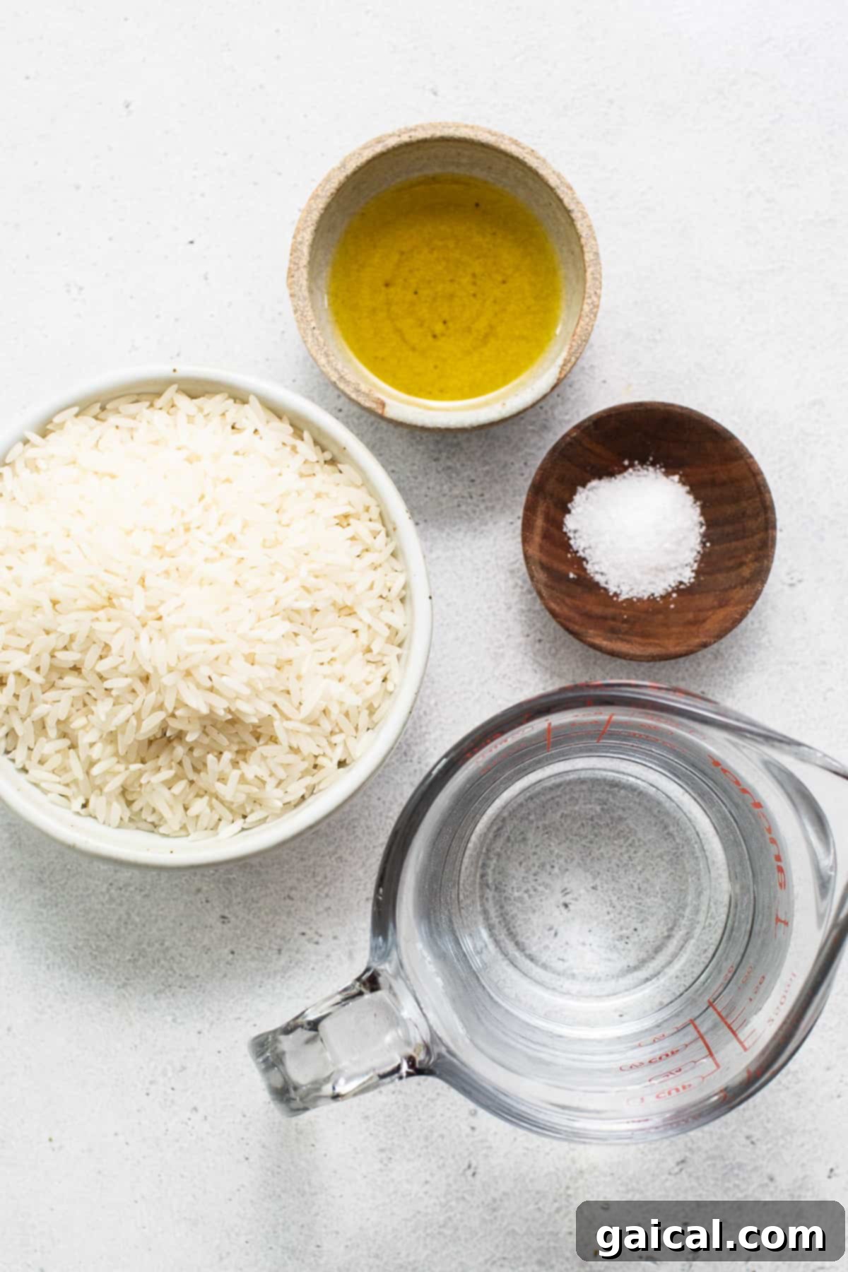A bowl of uncooked white rice, a bottle of olive oil, and a small dish of salt on a white surface.