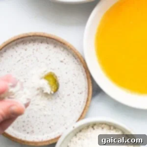 A hand gently sprinkling a generous amount of flour over a pickle slice, ensuring even coverage during the crucial breading process.