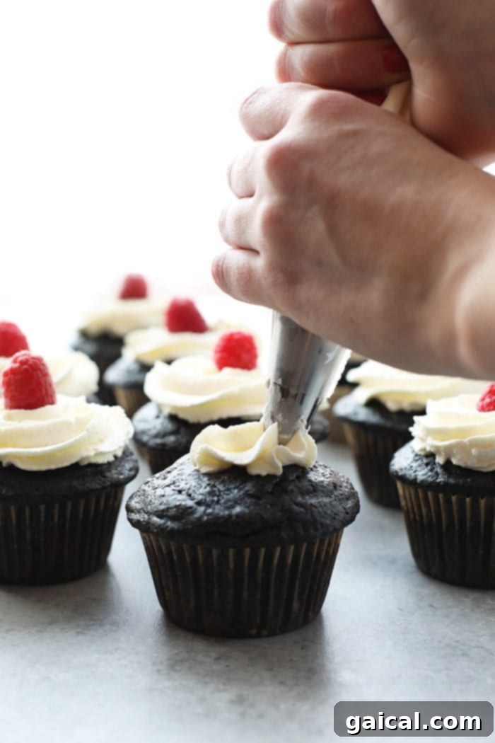 Healthier chocolate cupcakes being piped with creamy vanilla frosting and adorned with a fresh raspberry