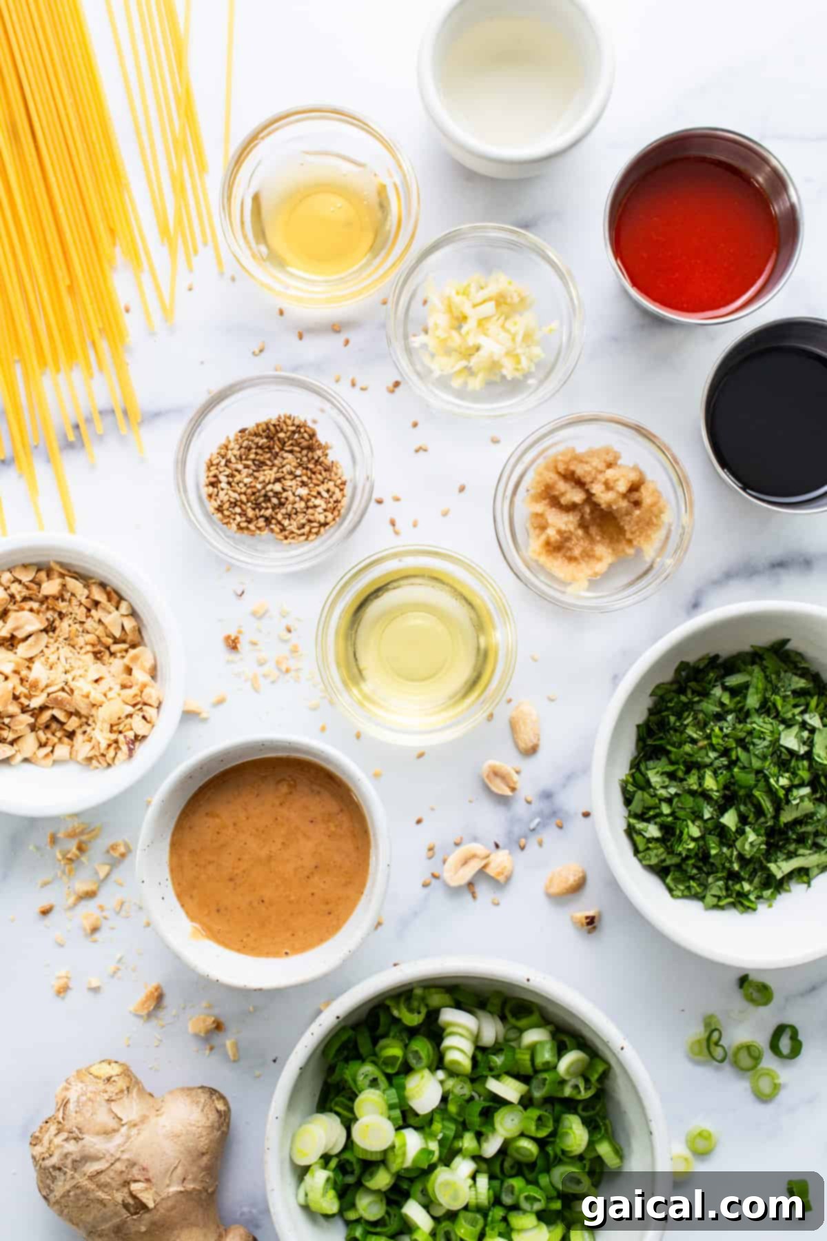 A vibrant selection of essential ingredients for making Asian-inspired noodles, including jars of peanut butter and soy sauce, bottles of sriracha and sesame oil, alongside fresh ginger, garlic, and rice vinegar, all neatly arranged on a white marble surface.