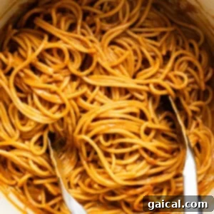 Freshly cooked spaghetti being generously tossed with a rich, creamy peanut sauce in a large blue pot using two forks, ensuring every strand is coated.