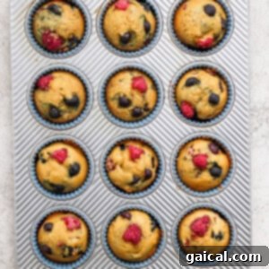 A muffin tin filled with golden-brown Greek yogurt berry muffins, gently cooling, with additional berries scattered artistically.