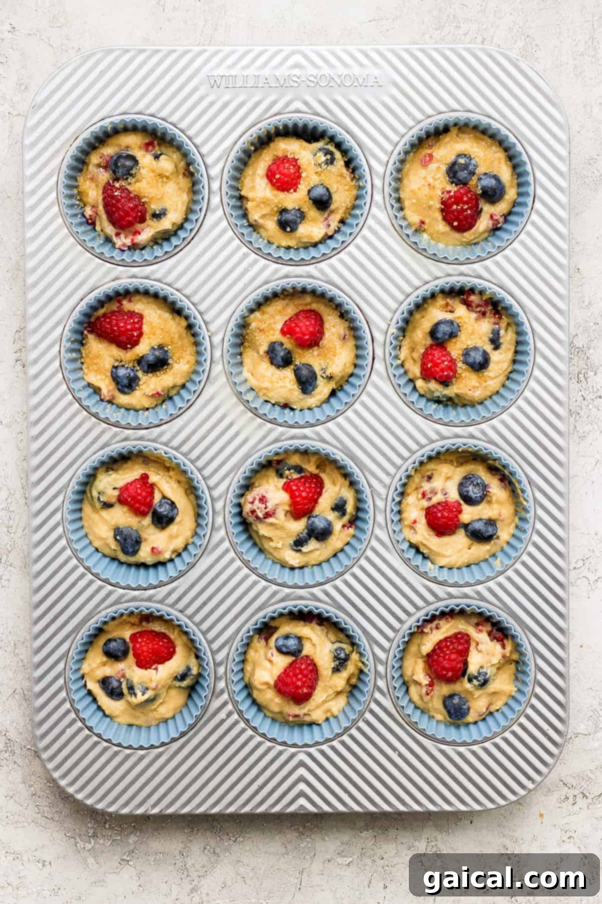 A standard muffin tin brimming with unbaked Greek yogurt berry muffin batter, topped with extra blueberries and raspberries, ready for the oven.