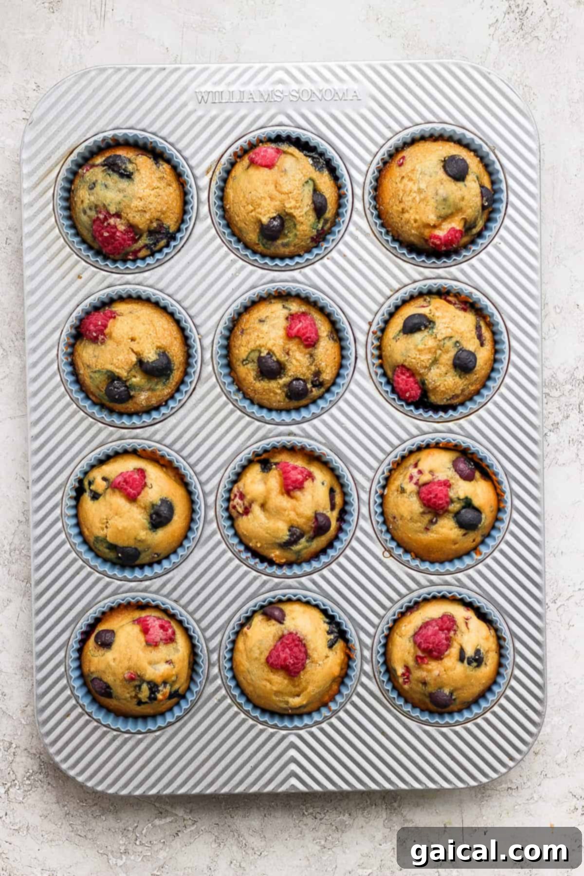 Freshly baked Greek yogurt berry muffins, still warm in the muffin tin, adorned with a scattering of vibrant raspberries and blueberries.