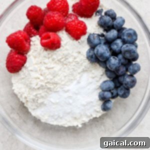 A bowl containing dry ingredients including flour and a colorful mix of blueberries and raspberries, prepped for muffin batter.