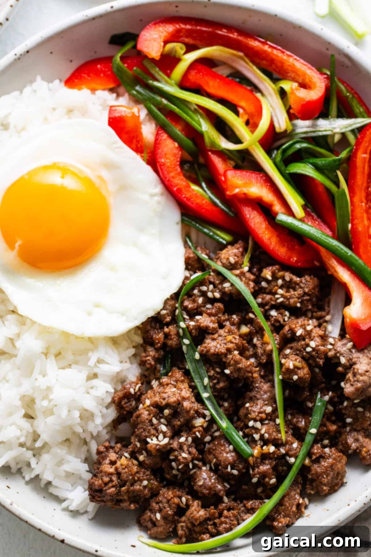 Sweet and Spicy Korean Ground Beef 2 A bowl of white rice topped with a sunny side up egg, seasoned Korean ground beef, sliced red bell peppers, and green onions, sprinkled with sesame seeds.